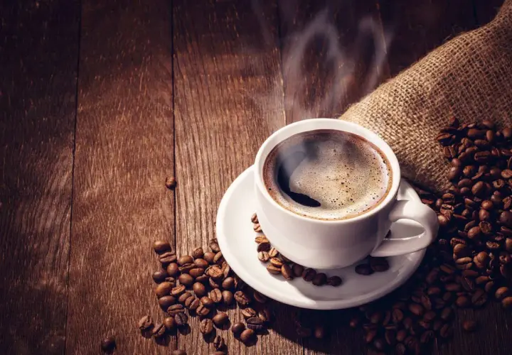 Tazza di caffè forte fumante su un piattino bianco, circondata da chicchi di caffè tostati su un tavolo di legno scuro.