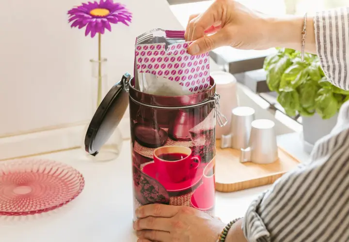 Mano inserisce pacchetto di caffè in una lattina decorata con chicchi e tazzina. Si può congelare il caffè liquido per conservarlo al meglio.