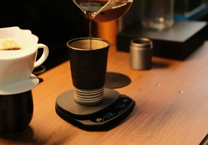 Versando caffè in una tazza scura, un'arte del coffee design. Bilancia digitale e filtro in primo piano.