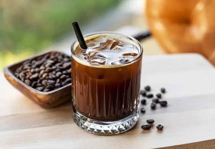 Un bicchiere di **cold brew latte** con ghiaccio e una cannuccia nera, accanto a una ciotola di chicchi di caffè tostati.