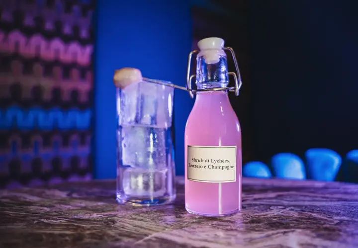 Bottiglia di shrub cocktail rosa con etichetta "Lychees, Zenzero e Champagne" accanto a un bicchiere pieno di ghiaccio.