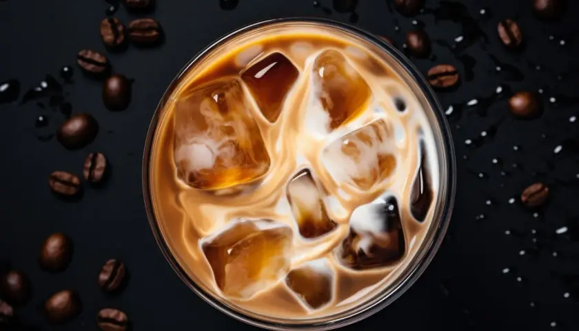 Un delizioso caffè freddo al bar, con cubetti di ghiaccio e latte che creano un vortice invitante. Chicchi di caffè tostati completano la scena.