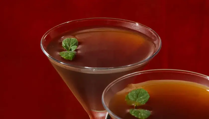 Due cocktail Stinger IBA, con liquore alla menta e brandy, guarniti con foglie di menta fresca, su sfondo rosso.