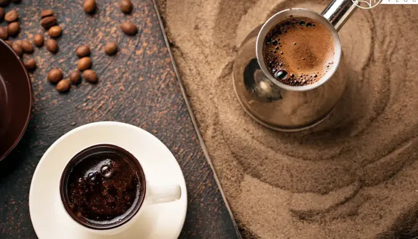 Un caffè arabo fumante in una tazzina bianca, accanto a un bricco di metallo su sabbia calda. Chicchi di caffè sparsi.