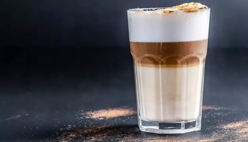 Un bicchiere di caffè macchiato caldo con strati di latte e caffè, sormontato da schiuma soffice e una spolverata di cacao.
