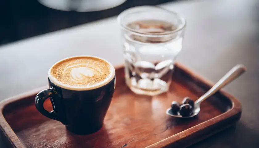 Tazza di caffè con latte art e bicchiere d'acqua, accompagnati da chicchi di caffè su un cucchiaino, su un vassoio di legno.