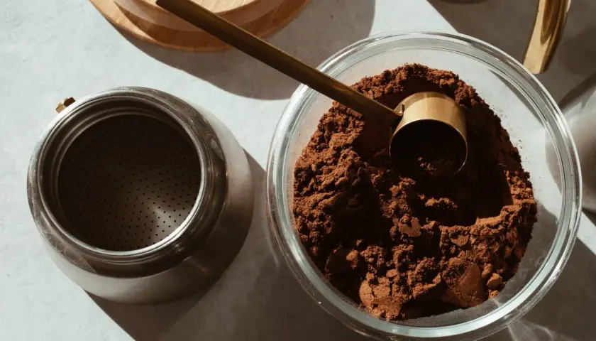 Caffè macinato in una ciotola di vetro, pronto per essere conservato. Un misurino dorato e una moka completano la scena, suggerendo come conservare il caffè al meglio.