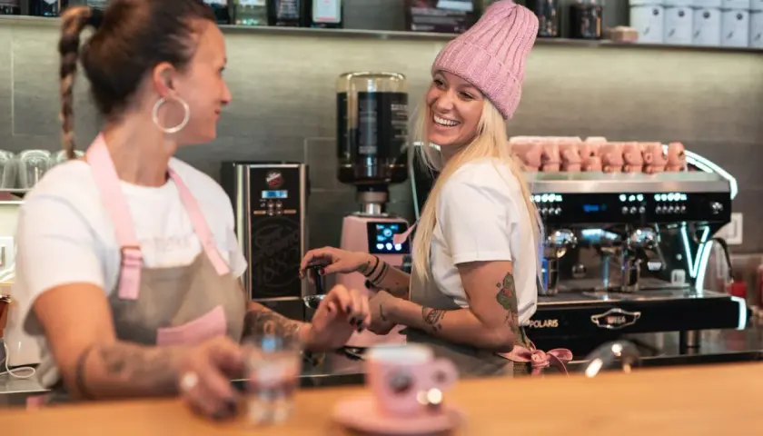 Due donne sorridono dietro il bancone di un bar, una con un cappello rosa, pronte a servire un caffè.