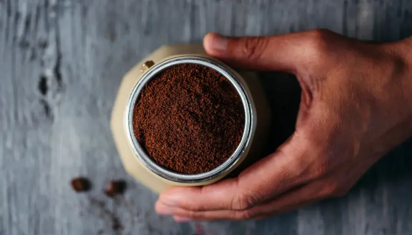 Mano che tiene la moka piena di caffè macinato, pronto per il rito del caffè. Chicchi sparsi sul tavolo di legno.