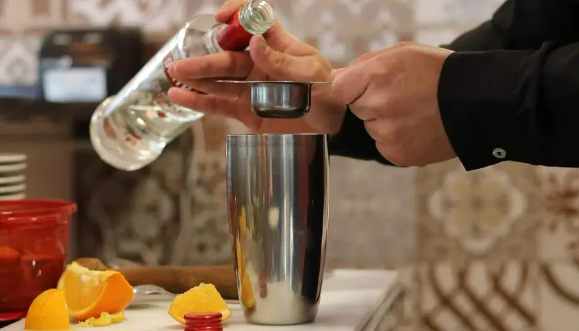 Preparazione di una ricetta cocktail: versando liquore da una bottiglia in un misurino sopra uno shaker, con spicchi d'arancia sullo sfondo.