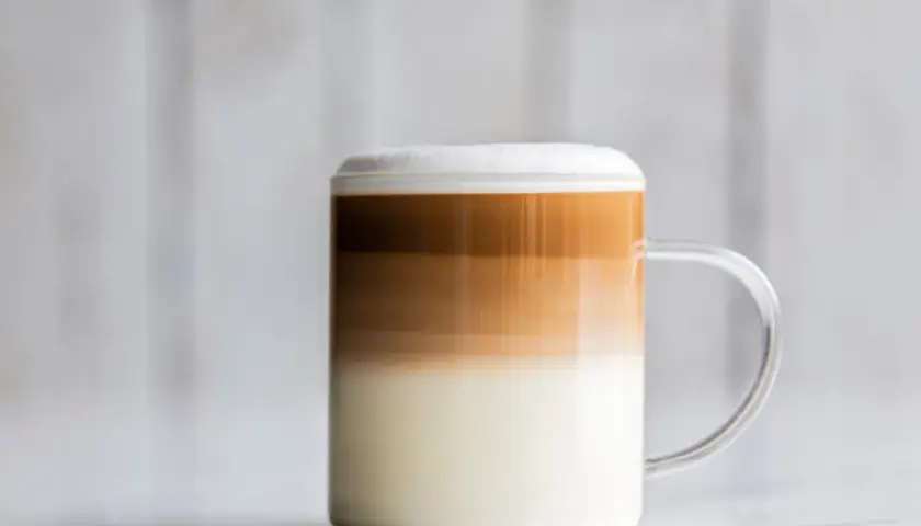 Un bicchiere di latte macchiato freddo con strati distinti di latte, caffè e schiuma, su uno sfondo sfocato.