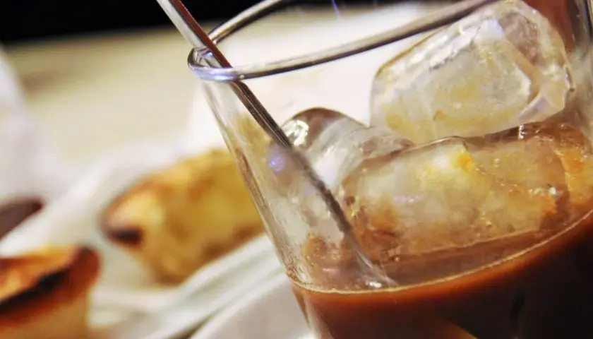 Un bicchiere di caffè con ghiaccio, perfetto per una pausa rinfrescante. Sullo sfondo, dolci sfocati invitano a un momento di piacere.