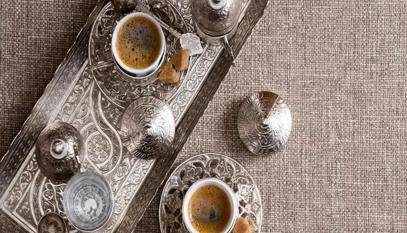 Un caffè in tazza grande, servito su un vassoio d'argento finemente decorato, con zollette di zucchero e bicchierino d'acqua.
