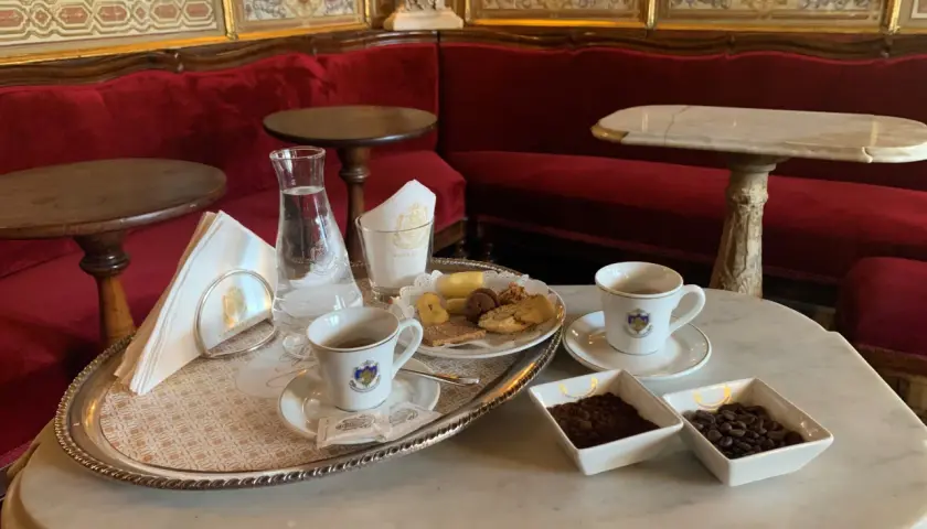 Un'elegante tazzina di caffè veneziano, biscotti e acqua su un vassoio d'argento, con divani rossi e decorazioni dorate sullo sfondo.