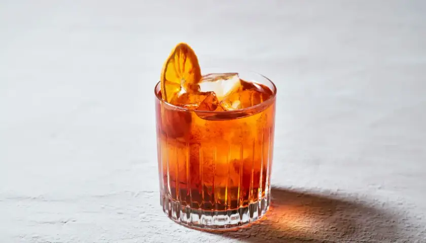 Un bicchiere di **bitter cocktail** arancione, con ghiaccio e una fetta d'arancia essiccata, su uno sfondo chiaro.