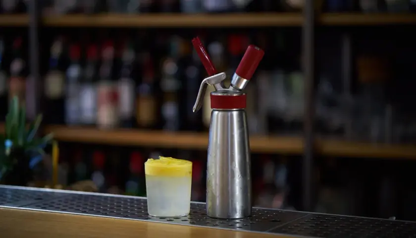 Sifone per panna montata accanto a un bicchiere con spuma drink giallo e bianco, su un bancone di bar con bottiglie sfocate sullo sfondo.
