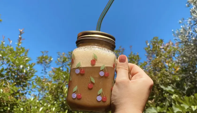 Un bicchiere di **iced coffee latte** decorato con ciliegie, tenuto in mano contro un cielo azzurro e alberi verdi.