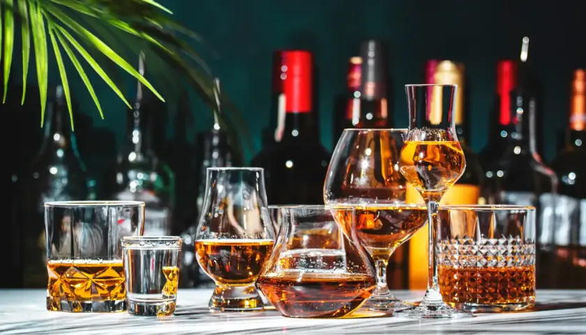 Varietà di bicchieri con ambra liquida, che evocano le bevande tipiche italiane. Sullo sfondo, bottiglie scure e una foglia verde.