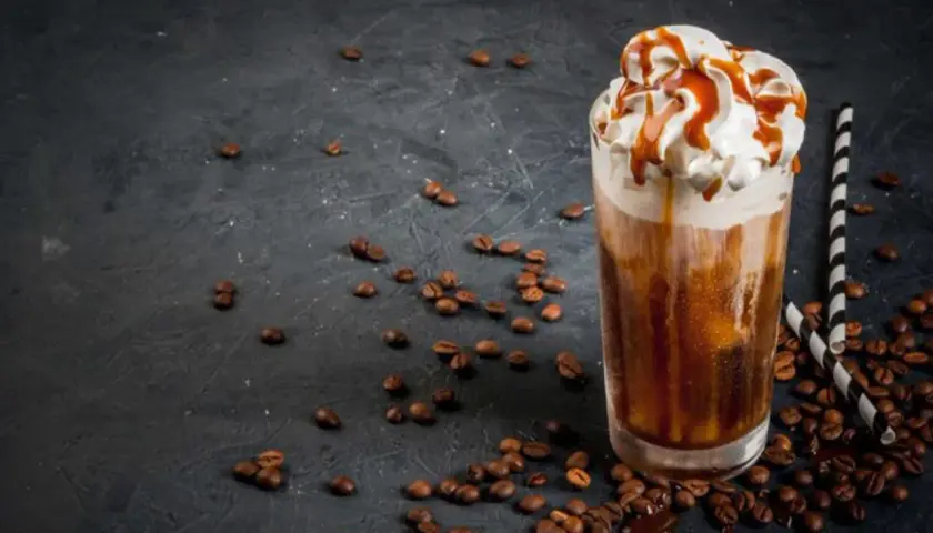 Un delizioso frappe al caffè con panna montata e sciroppo al caramello, circondato da chicchi di caffè tostati su sfondo scuro.