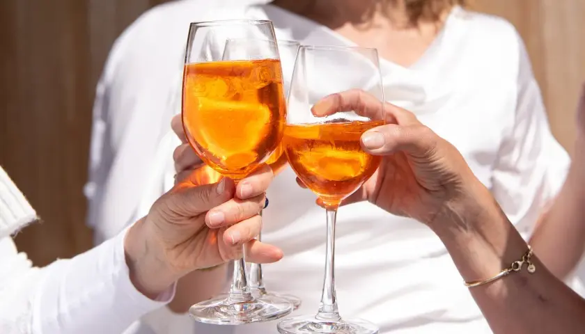 Tre mani brindano con bicchieri di Aperol, un drink rinfrescante e senza glutine, sotto il sole.