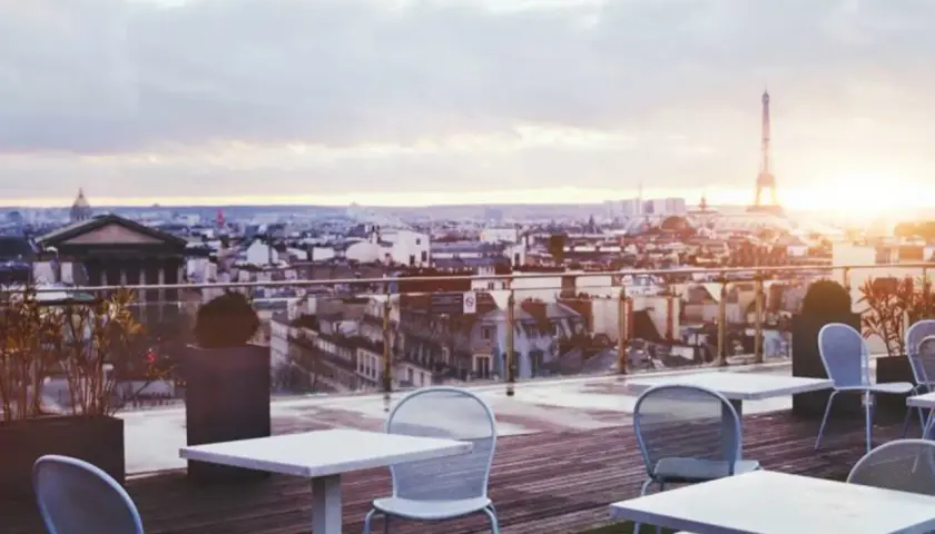Terrazza con vista su Parigi al tramonto, tavoli e sedie vuoti, la Torre Eiffel sullo sfondo. Un luogo perfetto per godersi i locali storici di Parigi.