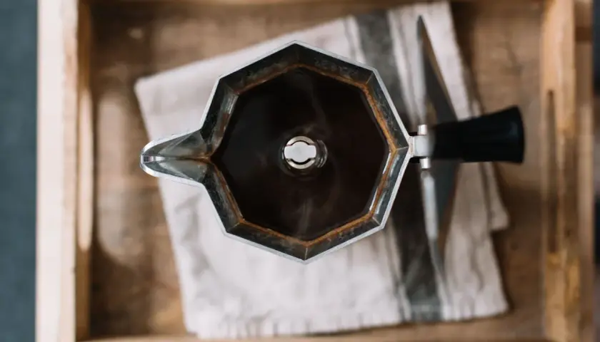 Moka nuova, appena usata, con caffè fumante, appoggiata su un panno a righe in una cassetta di legno.
