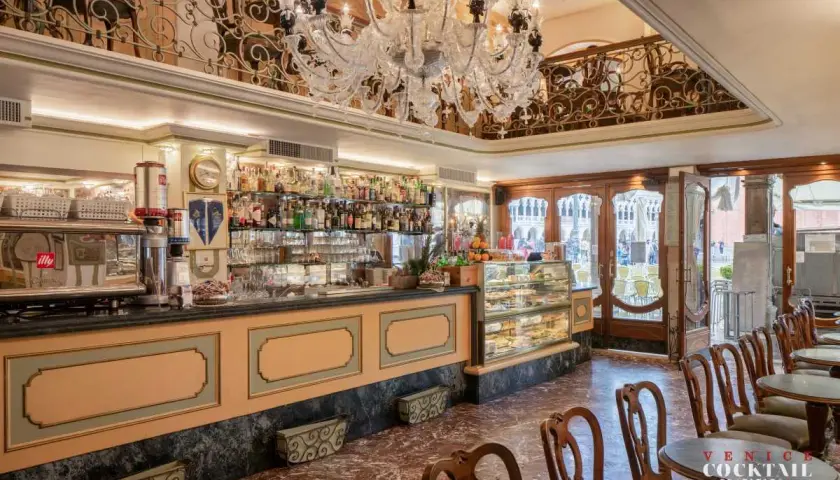 Interno di un caffè storico a Venezia, con bancone bar, vetrine di dolci, tavolini e un lampadario di cristallo.