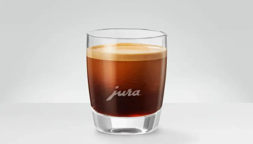 Un bicchiere di vetro con un doppio macchiato, la schiuma dorata corona il caffè scuro. Il logo Jura è inciso sul vetro.