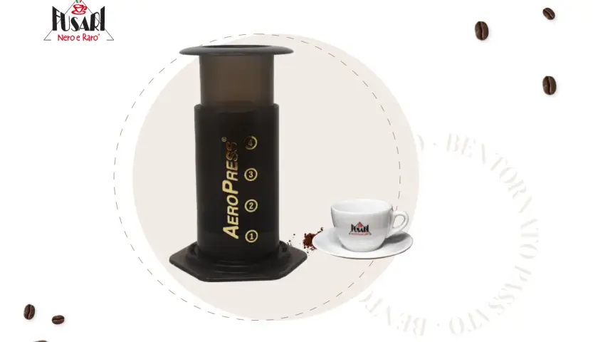 AeroPress per un caffè perfetto. Accanto, una tazzina bianca con logo e chicchi di caffè sparsi.