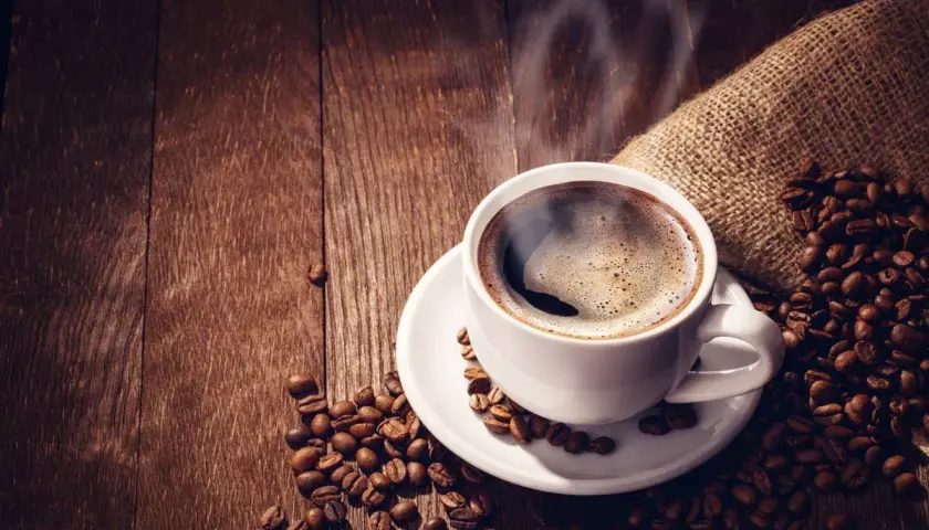 Tazza di caffè forte fumante su un piattino bianco, circondata da chicchi di caffè tostati su un tavolo di legno scuro.