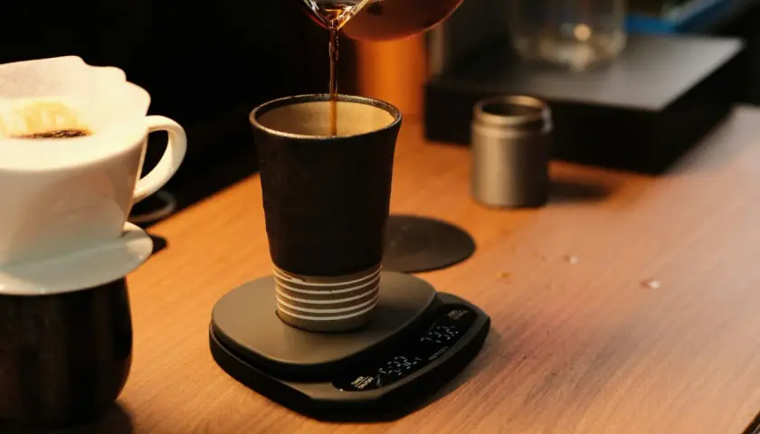Versando caffè in una tazza scura, un'arte del coffee design. Bilancia digitale e filtro in primo piano.