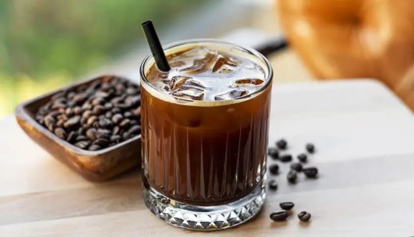 Un bicchiere di **cold brew latte** con ghiaccio e una cannuccia nera, accanto a una ciotola di chicchi di caffè tostati.