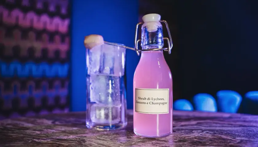 Bottiglia di shrub cocktail rosa con etichetta "Lychees, Zenzero e Champagne" accanto a un bicchiere pieno di ghiaccio.