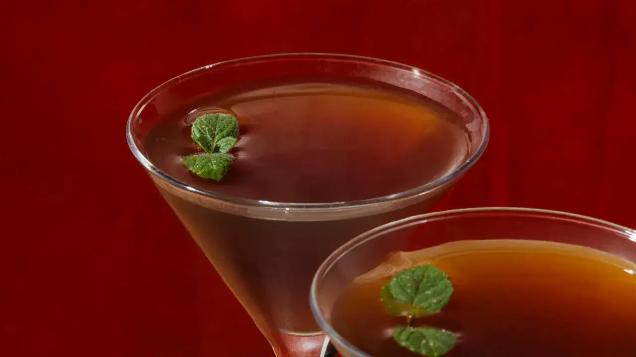 Due cocktail Stinger IBA, con liquore alla menta e brandy, guarniti con foglie di menta fresca, su sfondo rosso.