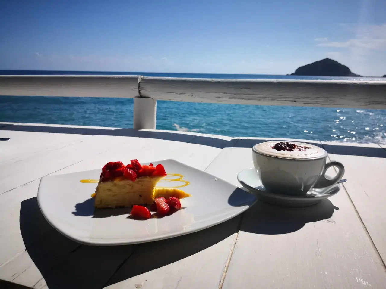 Un dolce con fragole e un caffè fumante, con il mare azzurro e un'isola all'orizzonte. Un momento di puro relax con un caffè al mare.