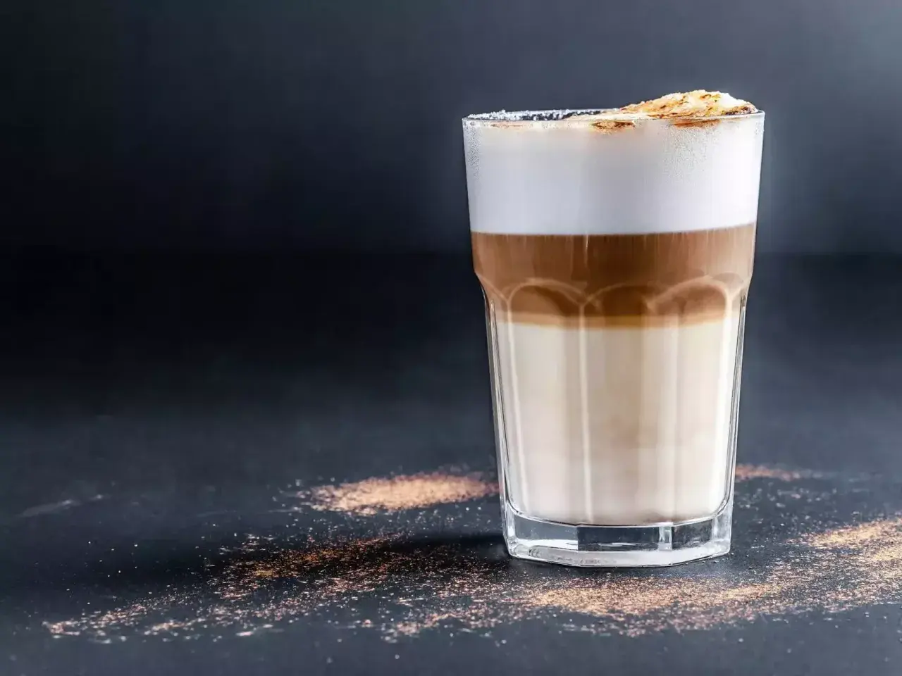 Un bicchiere di caffè macchiato caldo con strati di latte e caffè, sormontato da schiuma soffice e una spolverata di cacao.