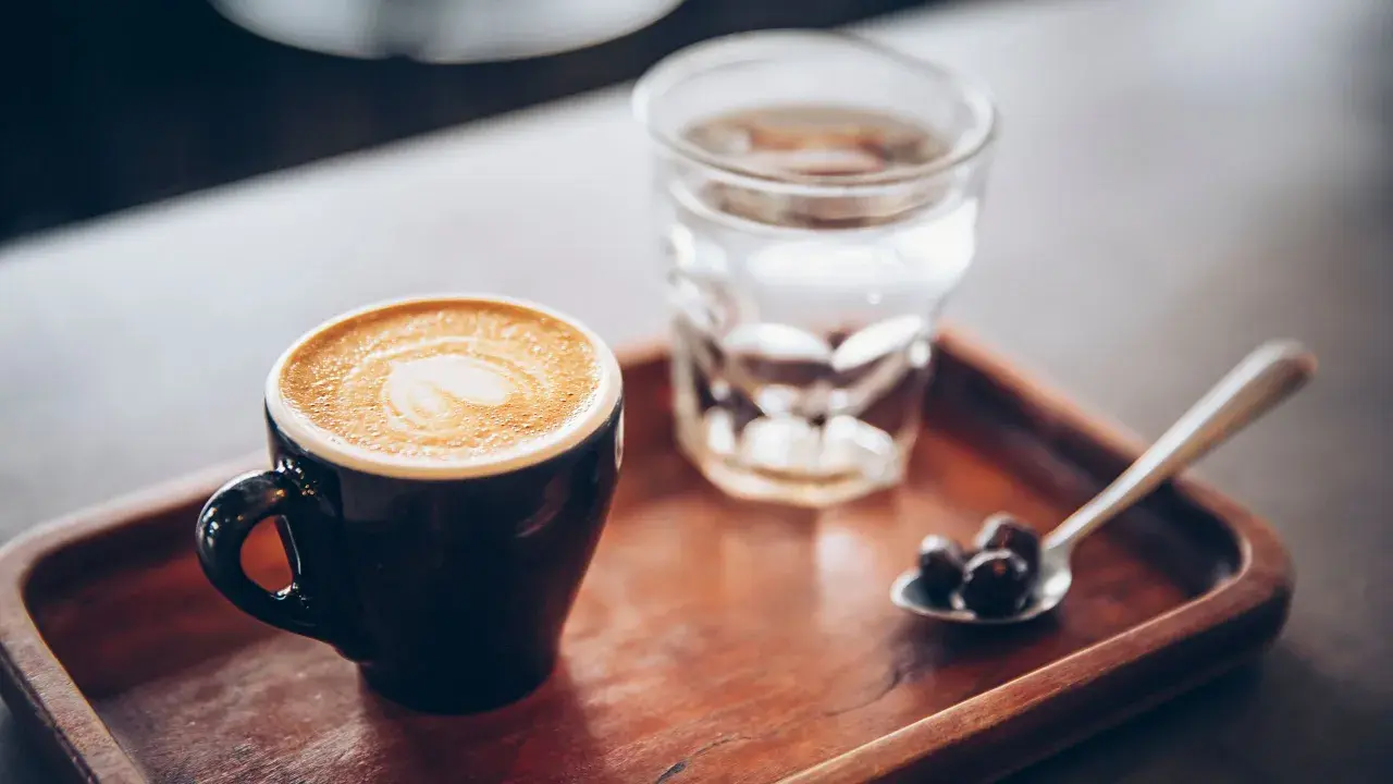 Tazza di caffè con latte art e bicchiere d'acqua, accompagnati da chicchi di caffè su un cucchiaino, su un vassoio di legno.