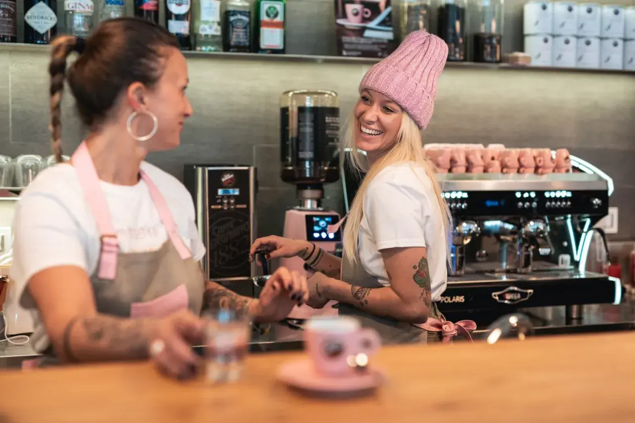 Due donne sorridono dietro il bancone di un bar, una con un cappello rosa, pronte a servire un caffè.