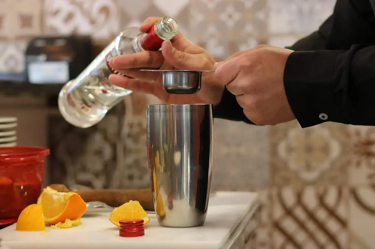 Preparazione di una ricetta cocktail: versando liquore da una bottiglia in un misurino sopra uno shaker, con spicchi d'arancia sullo sfondo.