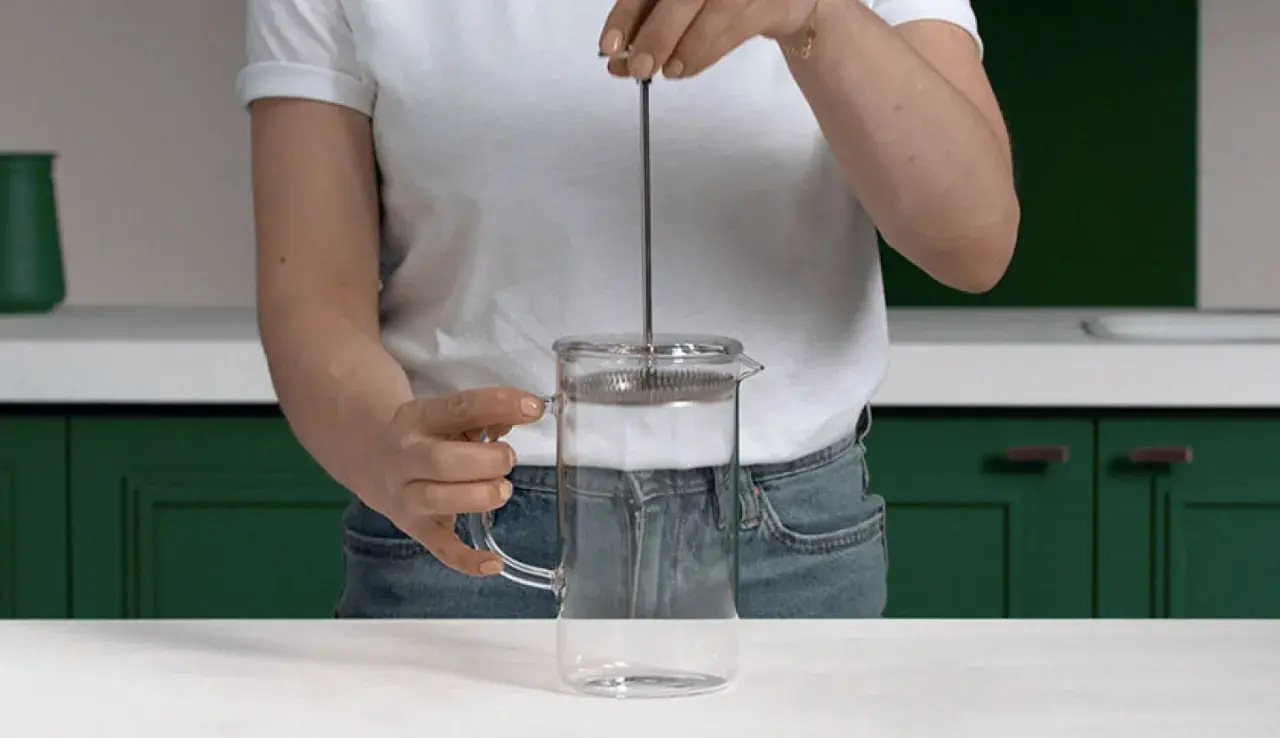 Mano che preme lo stantuffo di una french press di vetro, pronta per un caffè aromatico.