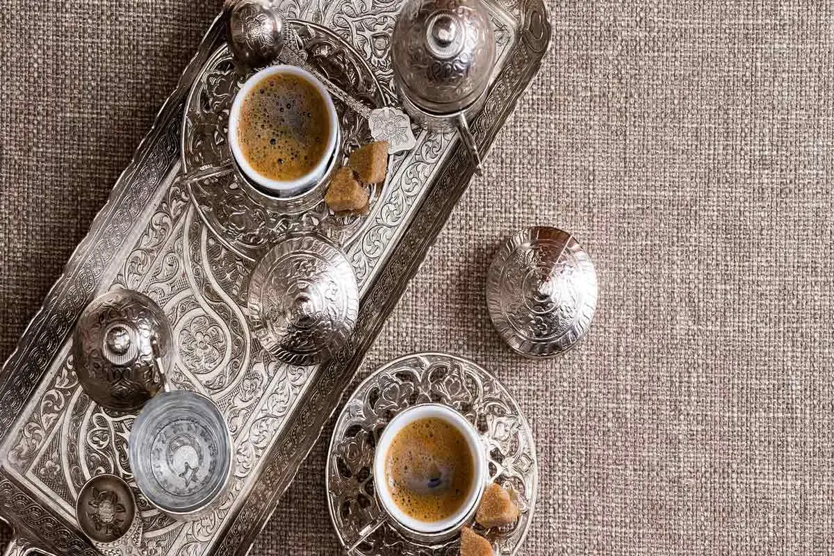 Un caffè in tazza grande, servito su un vassoio d'argento finemente decorato, con zollette di zucchero e bicchierino d'acqua.