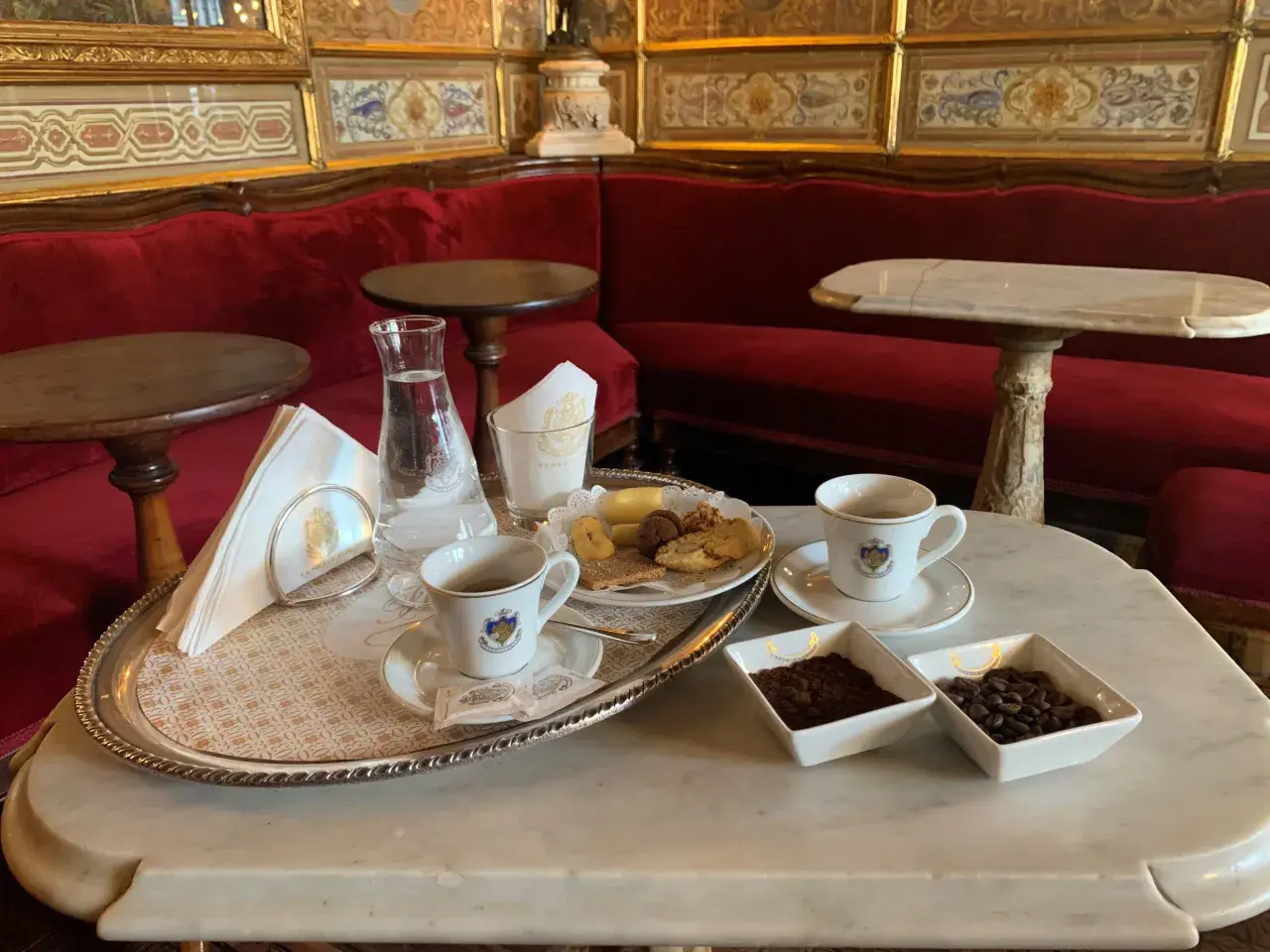 Un'elegante tazzina di caffè veneziano, biscotti e acqua su un vassoio d'argento, con divani rossi e decorazioni dorate sullo sfondo.
