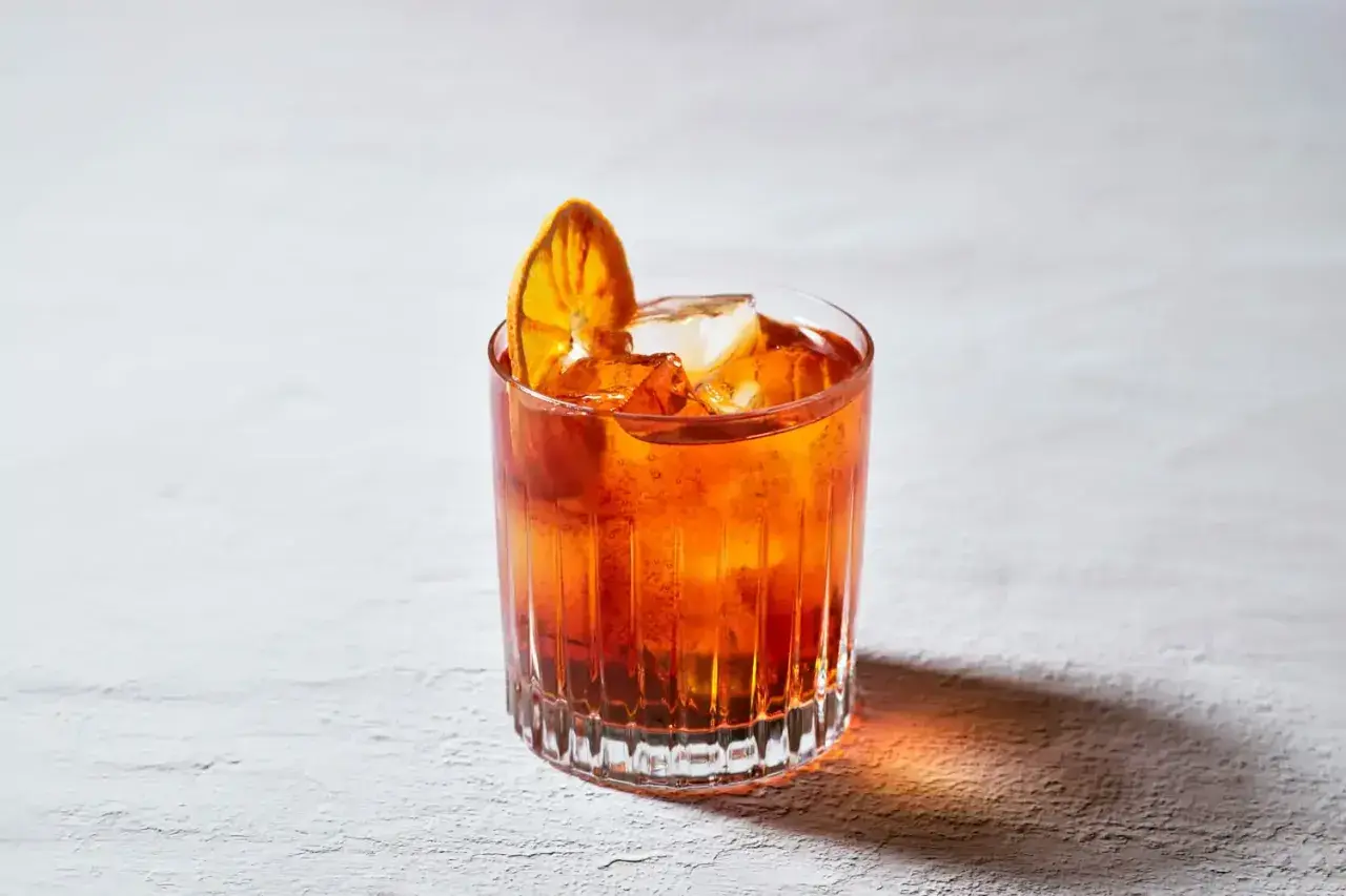 Un bicchiere di **bitter cocktail** arancione, con ghiaccio e una fetta d'arancia essiccata, su uno sfondo chiaro.