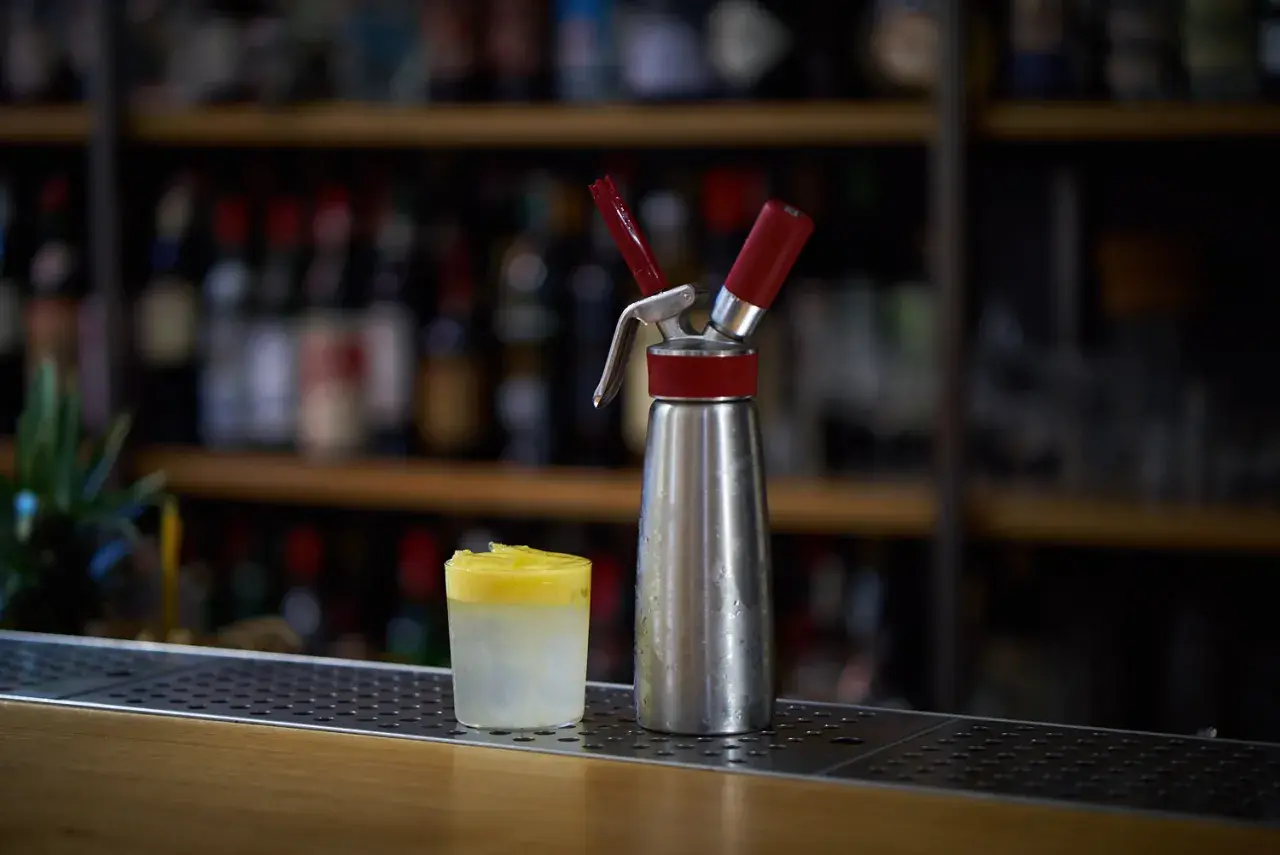 Sifone per panna montata accanto a un bicchiere con spuma drink giallo e bianco, su un bancone di bar con bottiglie sfocate sullo sfondo.