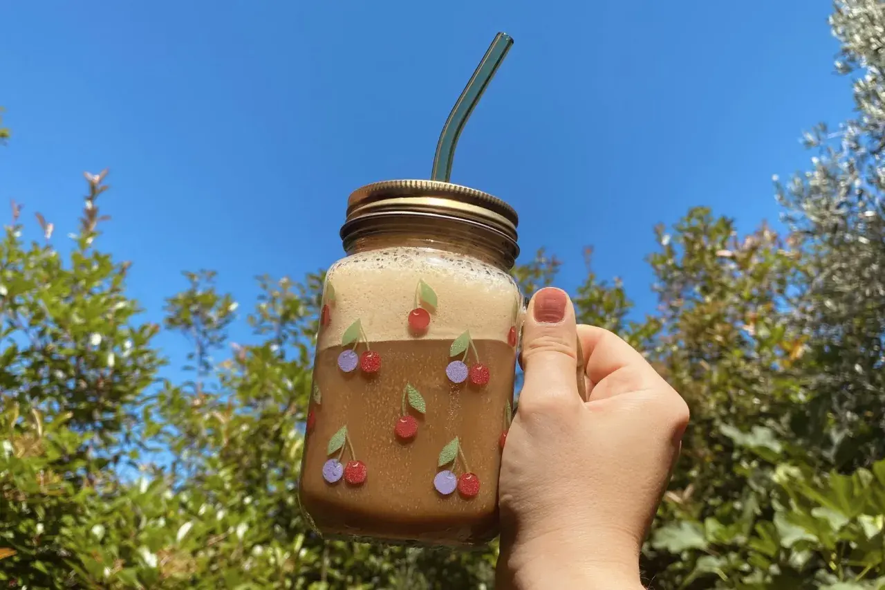Un bicchiere di **iced coffee latte** decorato con ciliegie, tenuto in mano contro un cielo azzurro e alberi verdi.