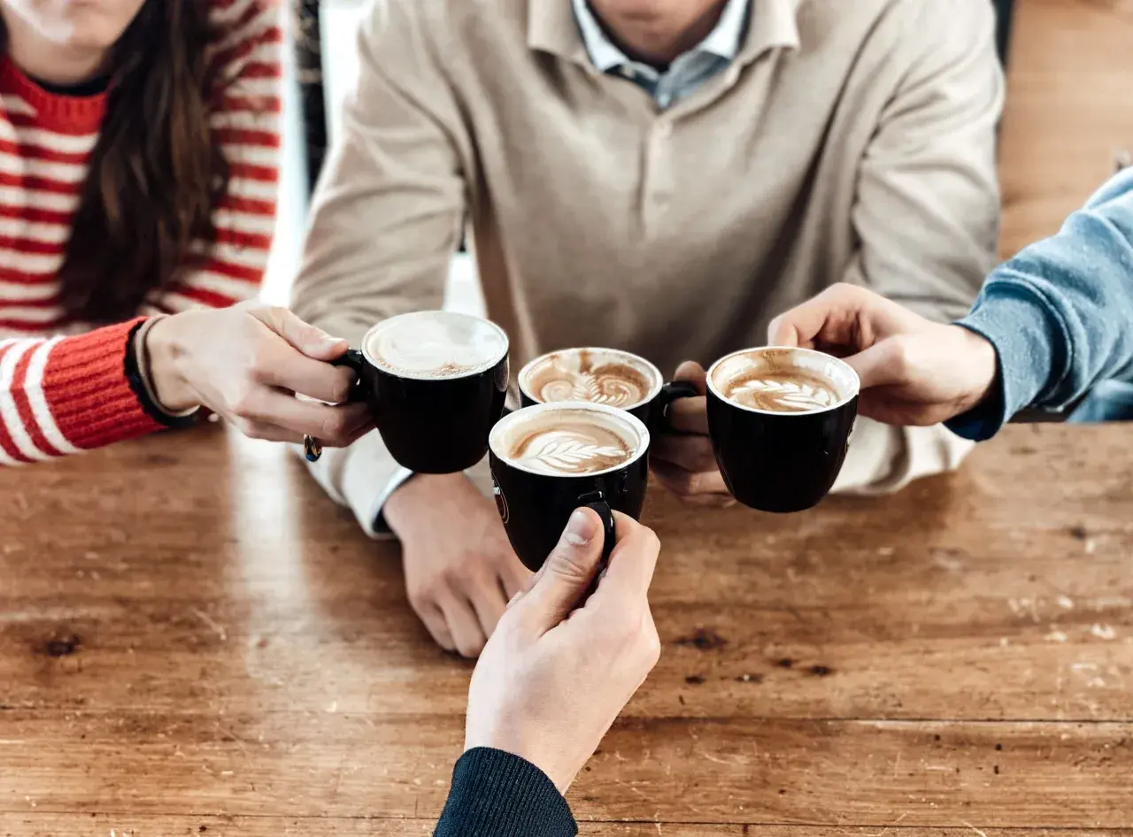 Un brindisi con caffè al bar con amici. Quattro tazze nere con schiuma artistica si avvicinano su un tavolo di legno.