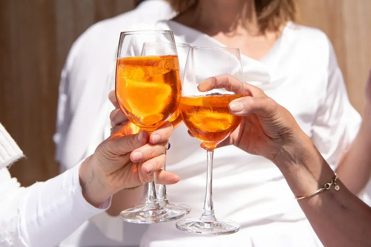 Tre mani brindano con bicchieri di Aperol, un drink rinfrescante e senza glutine, sotto il sole.