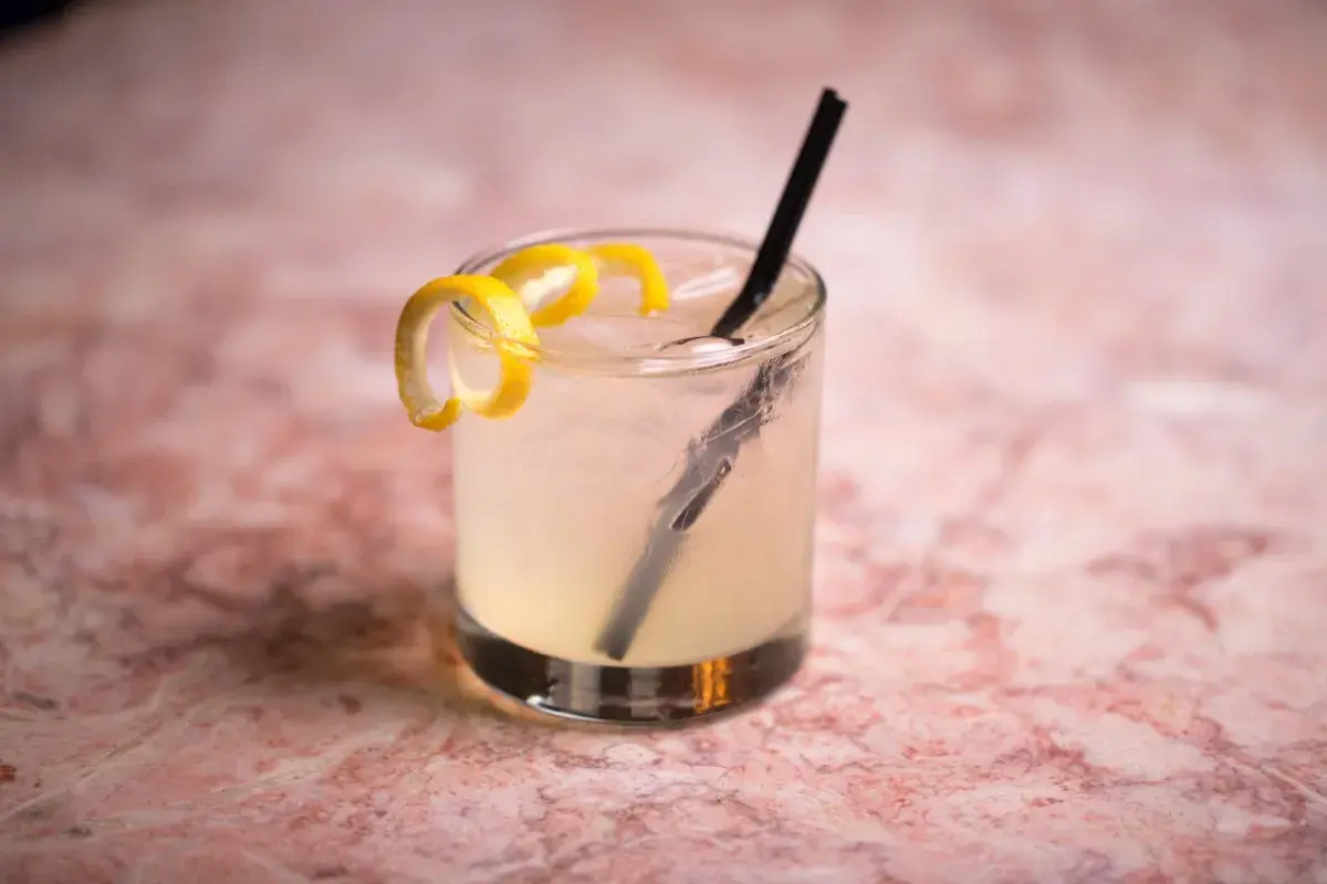 Un rinfrescante Vodka Sour IBA, guarnito con scorza di limone, su un tavolo di marmo rosa.