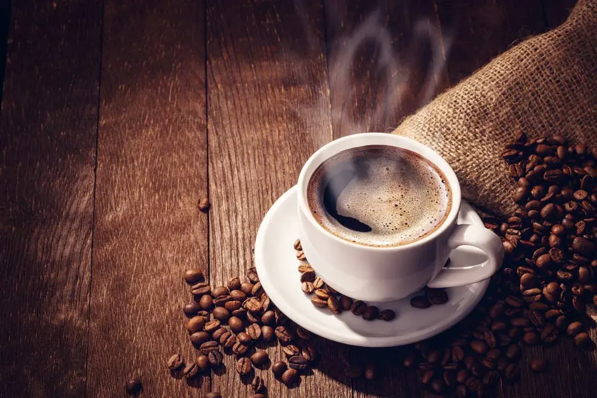 Tazza di caffè forte fumante su un piattino bianco, circondata da chicchi di caffè tostati su un tavolo di legno scuro.