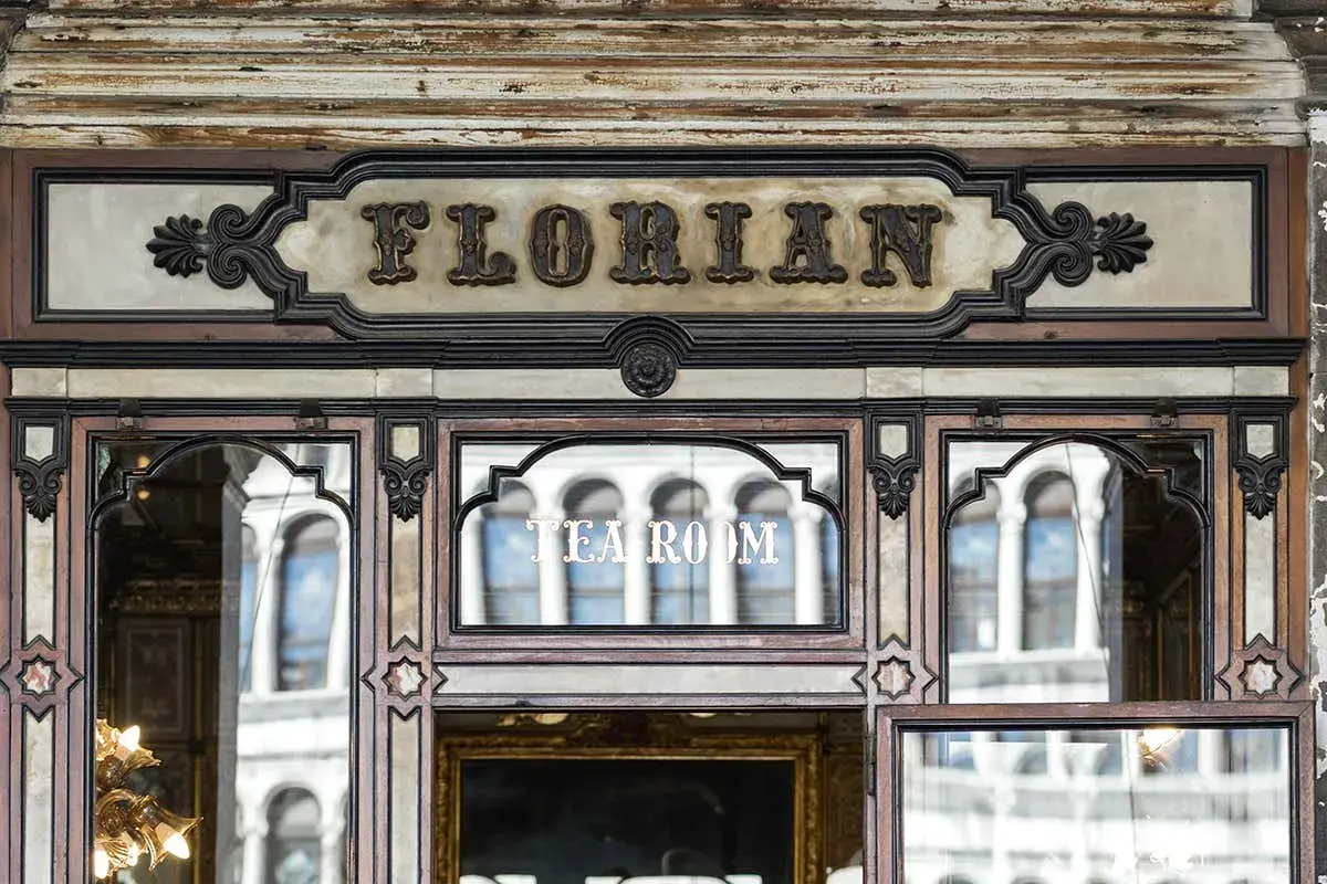 Ingresso del storico **Cafe Florian**, con insegna in rilievo e riflessi di palazzi veneziani nelle vetrate.